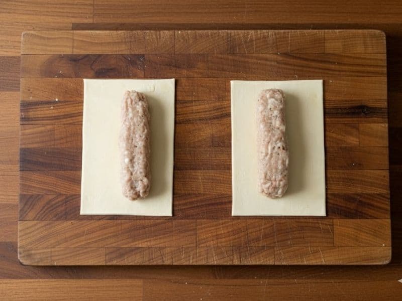 2 uncooked sausage rolls placed on a wooden chopping board on a wooden kitchen counter top.