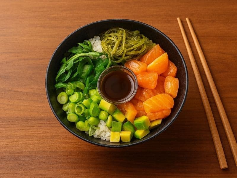 Yo! Sushi Salmon Poké Bowl — fresh salmon cubes served over sushi rice with avocado, seaweed salad, spring onions, and soy dressing in a black bowl with chopsticks on a wooden counter.