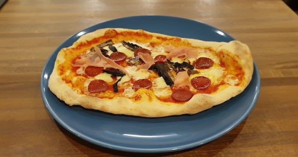Close-up of a Zizzi Pepperoni Campagna pizza topped with pepperoni, ham, and mushrooms, plated on a blue dish atop a wooden surface.