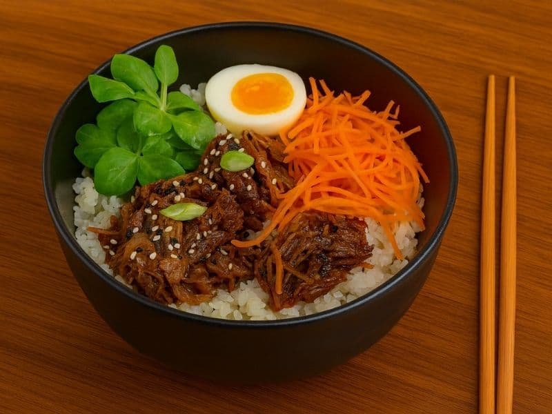 Wagamama-style beef donburi served in a black bowl with rice, shredded carrots, greens, sesame seeds, and a halved soft-boiled egg on a wooden countertop with chopsticks beside it.