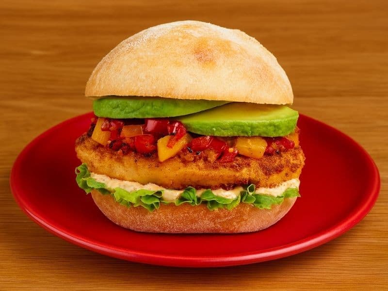 Close-up of Nando’s The Big Cheese burger on a red plate over a wooden countertop. The burger includes a crispy golden patty topped with diced red and yellow peppers, fresh avocado slices, lettuce, and creamy sauce in a soft ciabatta bun.