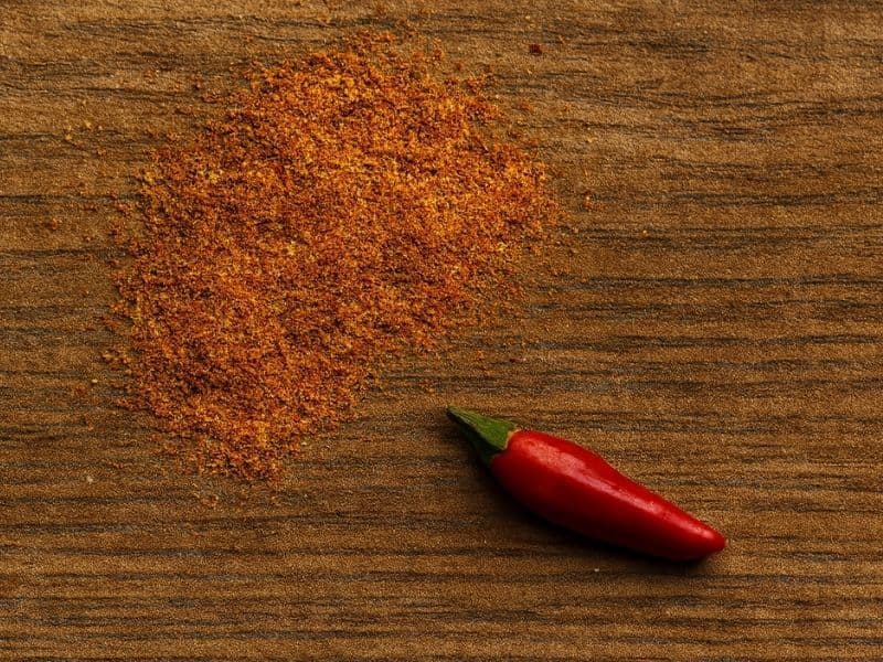 Close-up of a vibrant red peri-peri spice blend scattered on a rustic wooden table beside a single fresh red chilli pepper, captured in warm natural light.