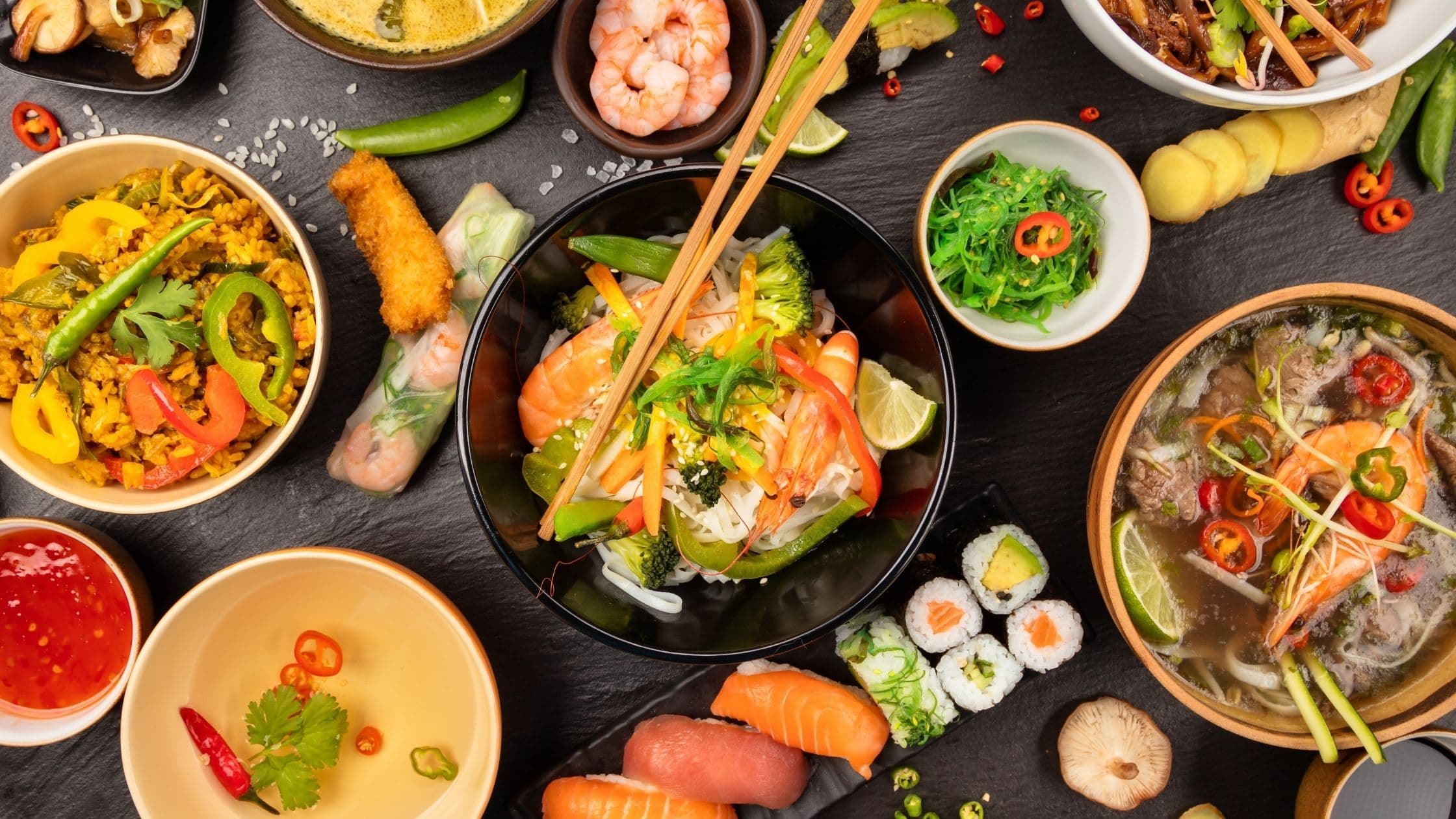 Overhead view of a colourful spread of Asian-inspired dishes on a dark slate surface, including noodle bowls, sushi rolls, prawns, spring rolls, fried rice, pho soup, and fresh vegetables with lime, chili, and herbs.