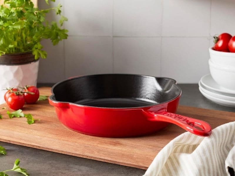 **Alt text:** Red STAUB 8.5-inch cast iron fry pan resting on a wooden kitchen countertop, surrounded by fresh tomatoes, parsley, and stacked white plates in a bright home kitchen setting.