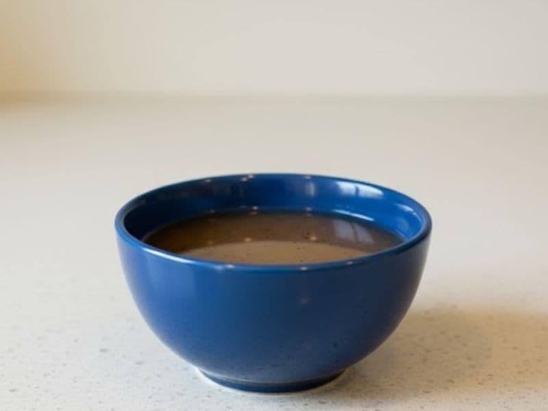 A small blue bowl filled with glossy brown sweet onion sauce, placed on a light speckled kitchen countertop. The sauce has a smooth texture and subtle sheen, captured in soft, natural indoor lighting.