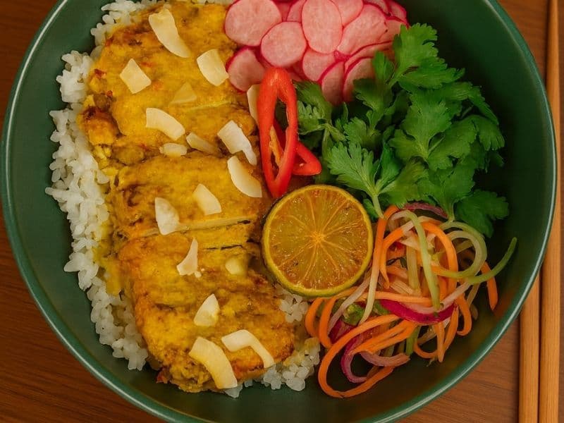 Wagamama-style Shu’s ‘Shiok’ Chicken served in a green bowl with rice, sliced radish, lime, coriander, and colourful vegetables on a wooden countertop with chopsticks beside it.