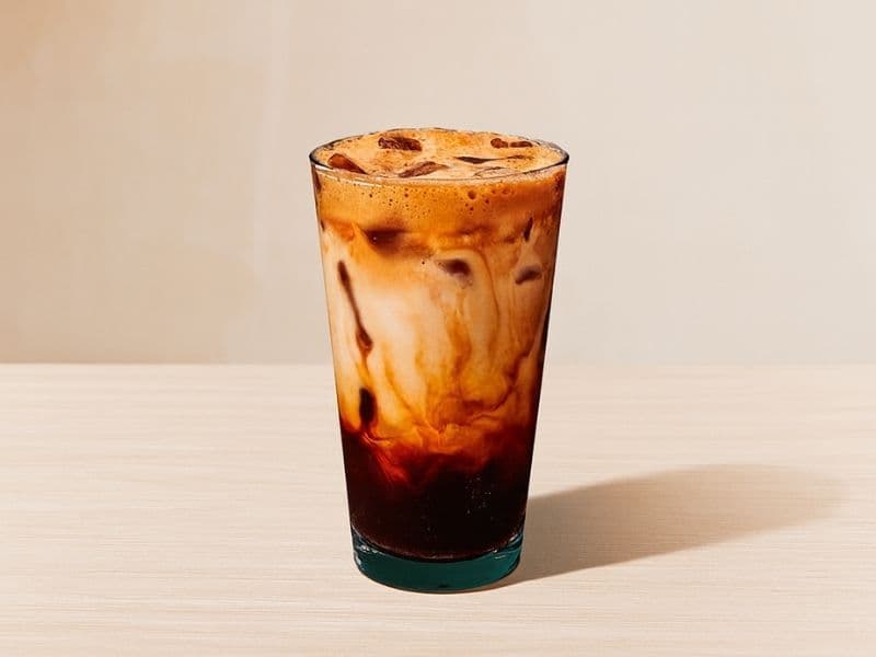 Tall glass of Starbucks-style iced brown sugar oat shaken espresso with layers of espresso and creamy oat milk swirling together over ice on a light wooden surface.