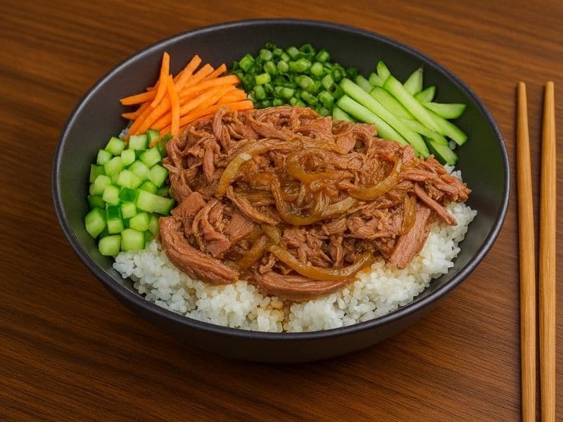 Wagamama-style duck donburi with shredded duck, rice, and fresh vegetables served in a black bowl on a wooden countertop with chopsticks beside it.