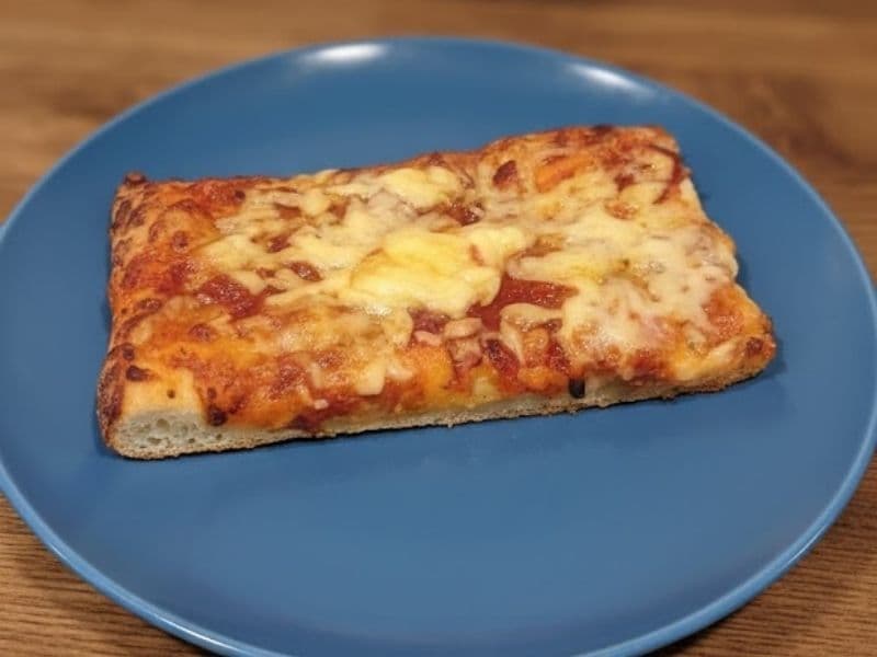 A rectangular slice of Greggs-style margherita pizza on a blue plate, featuring a golden, thick crust and melted cheese with tomato sauce on a wooden table background.