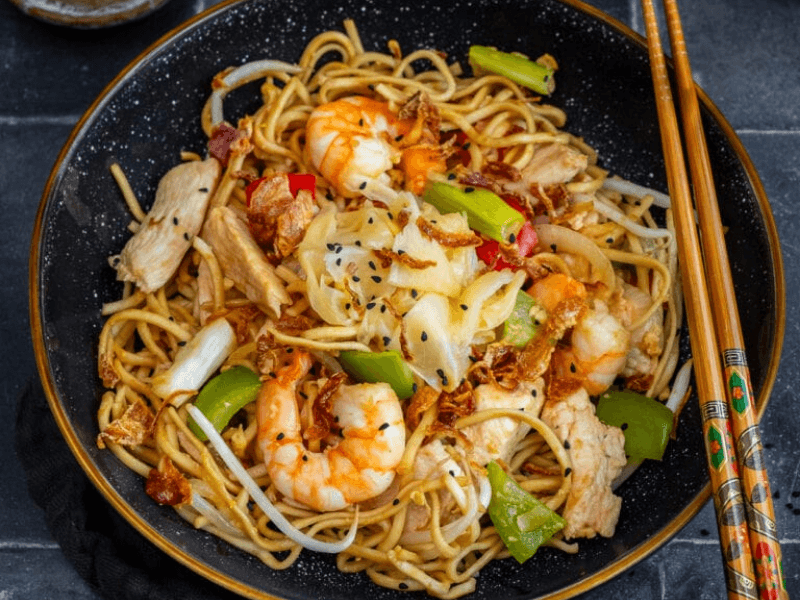 A black bowl of Wagamama-style Yaki Udon stir-fry with prawns, chicken, peppers, cabbage, bean sprouts, and sesame seeds, served with chopsticks.
