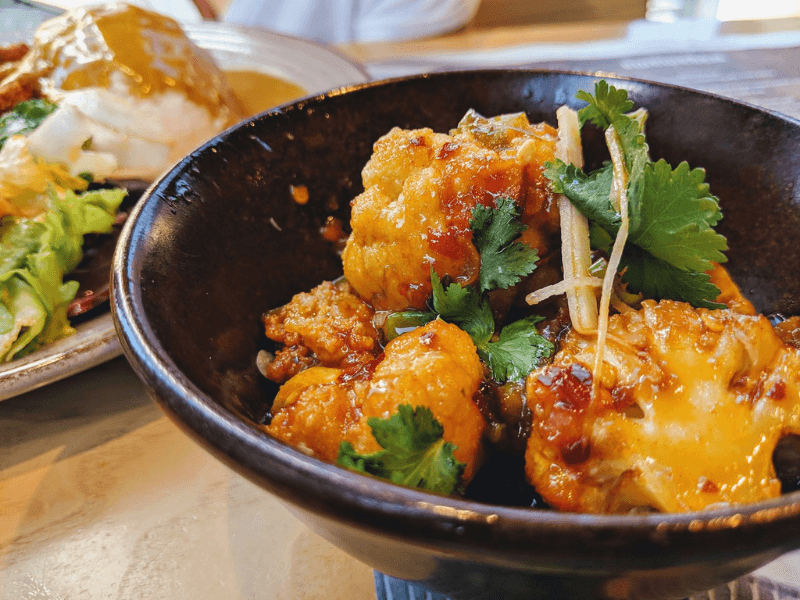 A black bowl of Wagamama-style Bang Bang Cauliflower, crispy golden cauliflower bites coated in a spicy-sweet sauce, garnished with fresh coriander and thin ginger strips.
