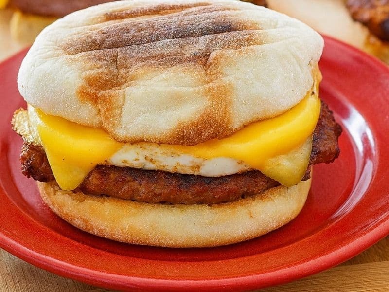 A homemade sausage and egg McMuffin sits on a bright red plate atop a wooden table. The sandwich features a toasted English muffin, melted cheese, fried egg, and a golden-brown sausage patty, with other similar breakfast sandwiches blurred in the background.