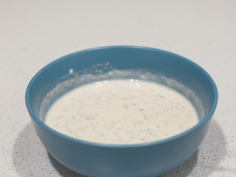 A light blue bowl filled with creamy white garlic and herb sauce, lightly speckled with herbs. The sauce has a smooth consistency and sits on a clean, speckled white countertop under soft indoor lighting.