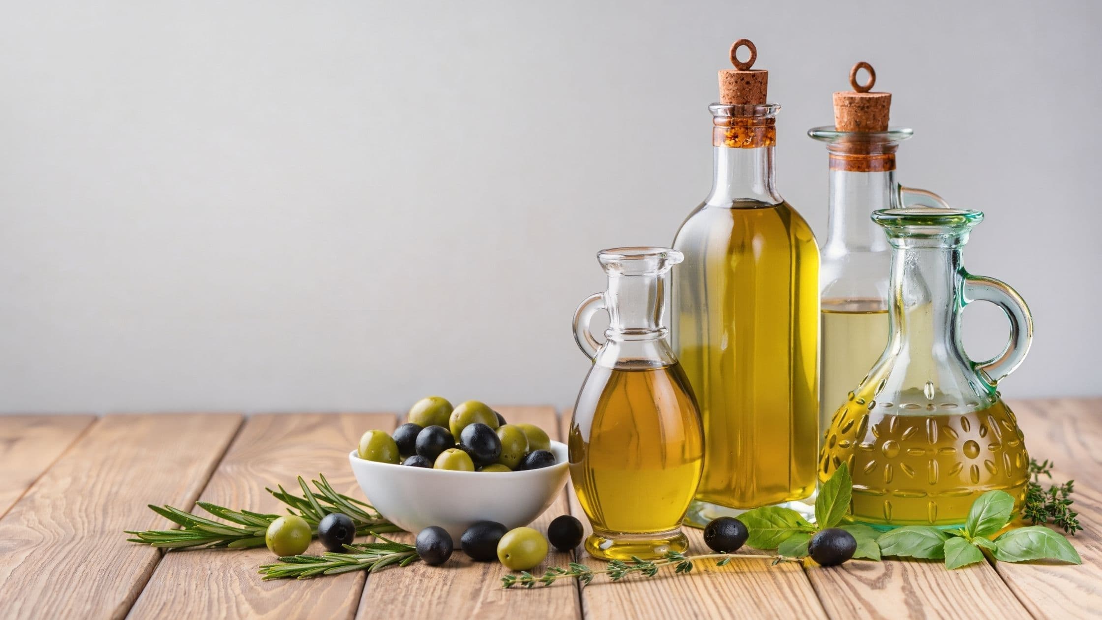 Different types of cooking oils in glass bottles with herbs and olives on a wooden counter.