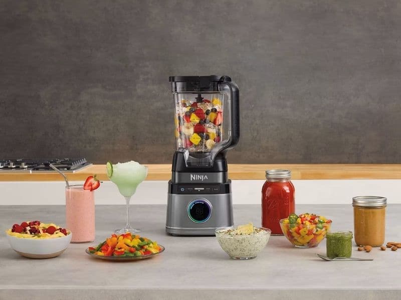 Alt text: A Ninja Detect Power Blender Pro 1200W (model TB201UK) sits on a modern kitchen countertop, filled with colorful chopped fruits. Around it are various blended foods and drinks — a strawberry smoothie, frozen cocktail, fruit salad, green pesto, red sauce, and nut butter — showcasing its versatility. The background is a clean grey kitchen wall with a stove in the distance.