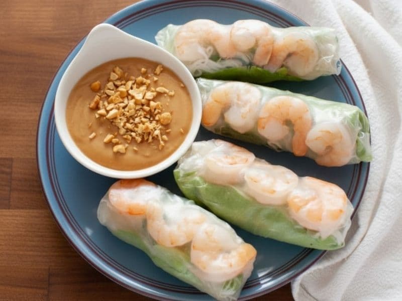 Four fresh Vietnamese summer rolls filled with prawns, lettuce, and rice noodles, served on a blue plate with a small bowl of creamy peanut dipping sauce topped with crushed peanuts.