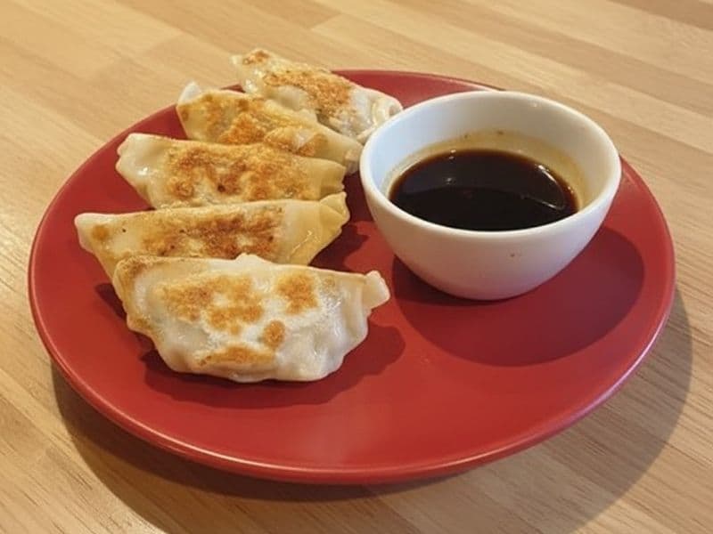 Pan-fried Wagamama chicken gyoza arranged on a red plate with a small white bowl of soy dipping sauce, set on a light wooden table.