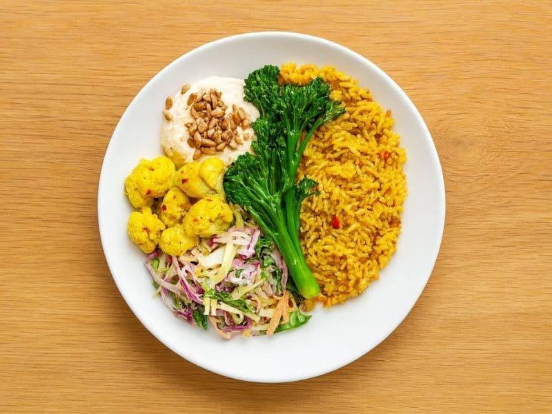 A white plate on a wooden tabletop filled with yellow spiced rice, tenderstem broccoli, golden cauliflower, creamy yogurt topped with sunflower seeds, and colourful cabbage slaw.