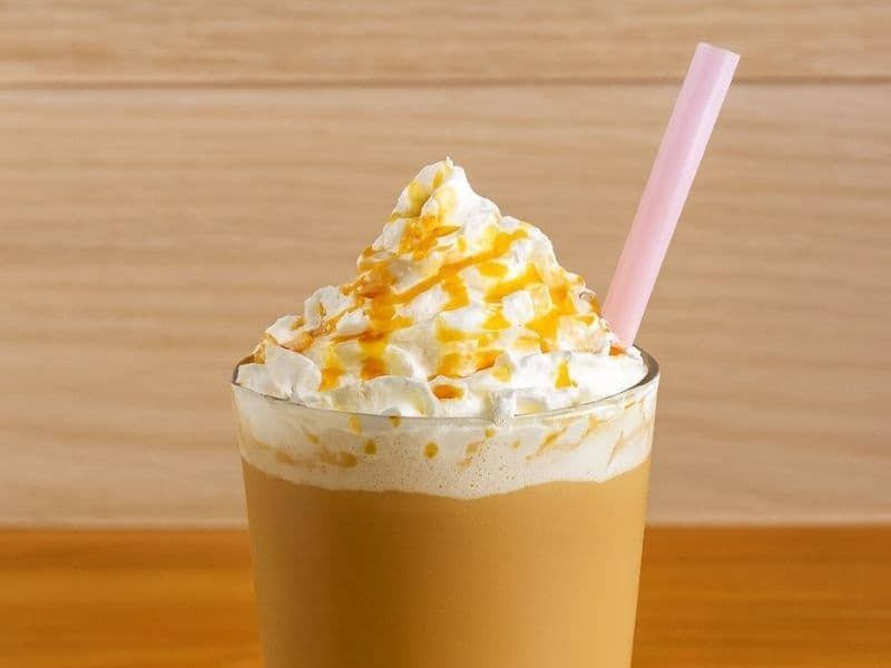 Starbucks-style iced caramel latte topped with whipped cream and caramel drizzle, served in a tall glass with a pink straw on a wooden countertop.