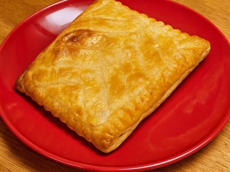 Golden-brown Greggs-style cheese and onion bake on a bright red plate, resting on a wooden kitchen countertop.